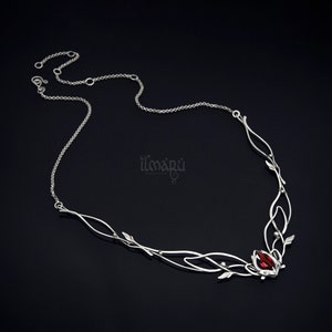 May include: A silver-toned necklace with a delicate, flowing design. The necklace features a central cluster with a vibrant red gemstone, accented by leaf-like details and a fine chain. The design is elegant and nature-inspired.