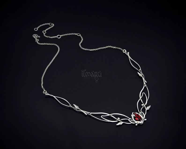 May include: A silver-toned necklace with a delicate, flowing design. The necklace features a central cluster with a vibrant red gemstone, accented by leaf-like details and a fine chain. The design is elegant and nature-inspired.