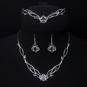 May include: A silver-toned jewelry set featuring a tiara, necklace, and earrings. Each piece has an intricate, flowing design with a central purple gemstone. The set is displayed against a black background, highlighting the delicate craftsmanship and elegant style.