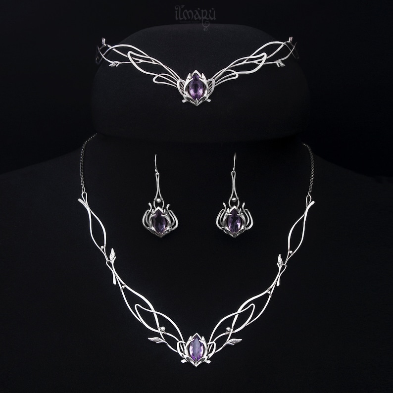 May include: A silver-toned jewelry set featuring a tiara, necklace, and earrings. Each piece has an intricate, flowing design with a central purple gemstone. The set is displayed against a black background, highlighting the delicate craftsmanship and elegant style.
