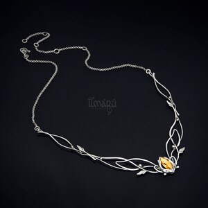 May include: A silver-toned necklace with an intricate, nature-inspired design. The necklace features delicate leaf and vine details, culminating in a central cluster with a yellow-orange gemstone. The necklace is displayed against a dark background.