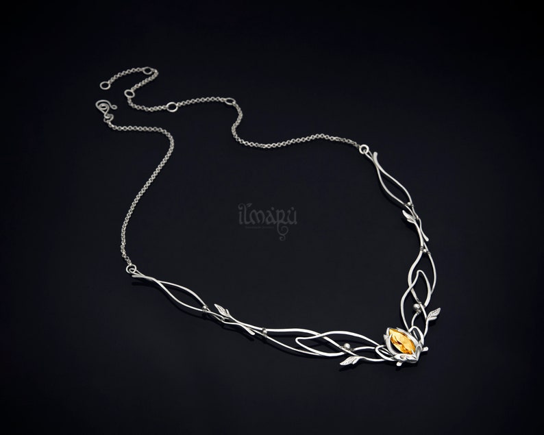 May include: A silver-toned necklace with an intricate, nature-inspired design. The necklace features delicate leaf and vine details, culminating in a central cluster with a yellow-orange gemstone. The necklace is displayed against a dark background.