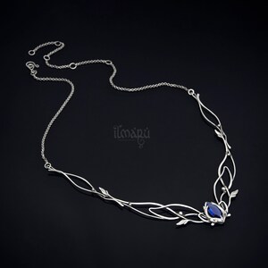 May include: A silver-toned necklace with an intricate, flowing design. The necklace features delicate leaf accents and a central, deep blue gemstone. The chain is fine and adjustable, with the brand name "ilmapu" visible on the right side.