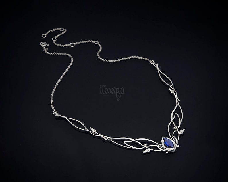May include: A silver-toned necklace with an intricate, flowing design. The necklace features delicate leaf accents and a central, deep blue gemstone. The chain is fine and adjustable, with the brand name "ilmapu" visible on the right side.