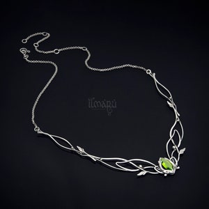 May include: A silver-toned necklace with a leaf and vine design. The necklace features a central green gemstone and delicate chain. The design includes small leaf accents and the brand name "ilmapu".