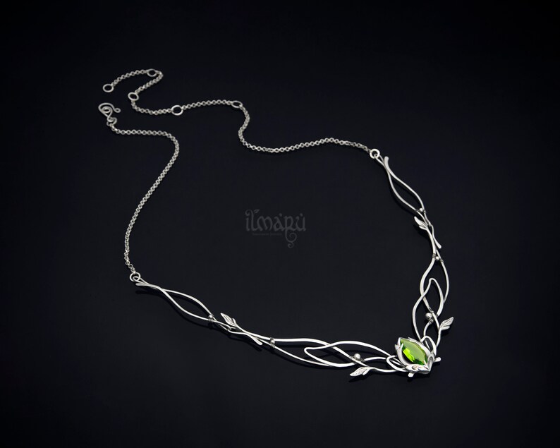 May include: A silver-toned necklace with a leaf and vine design. The necklace features a central green gemstone and delicate chain. The design includes small leaf accents and the brand name "ilmapu".