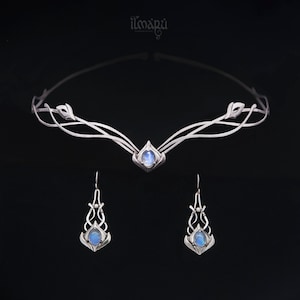 May include: A silver-toned jewelry set featuring a delicate tiara and matching earrings. The tiara and earrings have an elegant, flowing design with leaf-like accents and a central, oval-shaped, light blue stone. The brand name "ilmapü" is at the top.