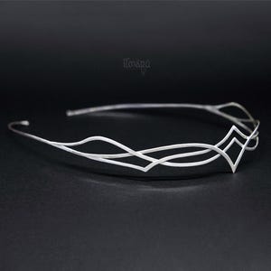 May include: A delicate silver-toned circlet crown with an intricate, flowing design. The crown features a series of interconnected wavy lines and diamond-shaped accents. The crown is displayed against a dark background, highlighting its elegant details.