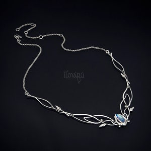 May include: A silver necklace with an intricate design featuring delicate leaves and flowing lines. The necklace has a central gemstone and is displayed against a dark background. The brand name "ilmapu" is visible.