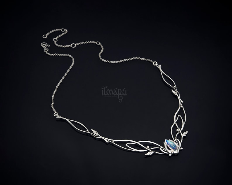 May include: A silver necklace with an intricate design featuring delicate leaves and flowing lines. The necklace has a central gemstone and is displayed against a dark background. The brand name "ilmapu" is visible.