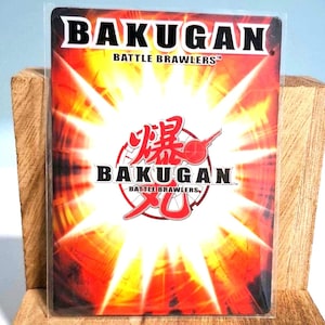 Peut inclure: Une carte à collectionner Bakugan Battle Brawlers. La carte présente un fond rouge et jaune avec le mot "BAKUGAN" en blanc en haut et en bas. La carte comporte également des caractères japonais et les mots "Battle Brawlers" en blanc.