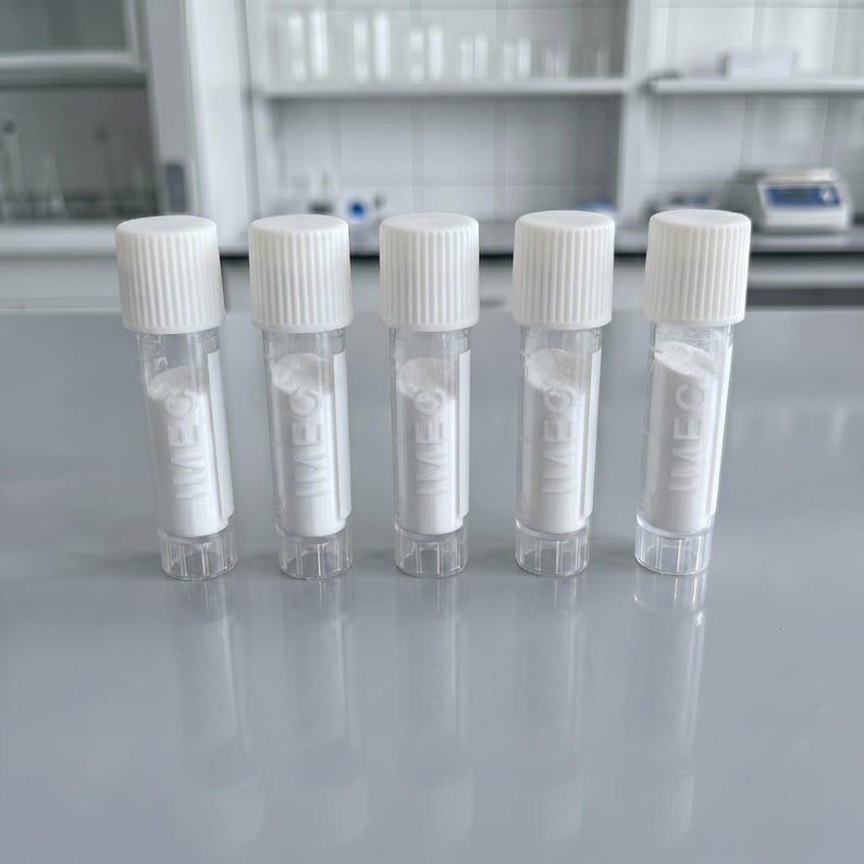 Pu&ograve; includere: Cinque piccoli flaconi trasparenti con tappi a vite bianchi. Ogni flacone contiene una polvere bianca e riporta la scritta "IMEC". I flaconi sono disposti in fila su una superficie grigio chiaro, probabilmente in un laboratorio.