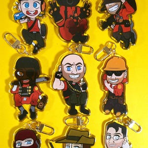 May include: Nine acrylic keyrings featuring cartoon characters in various poses. The characters are dressed in red, black, and brown outfits, with some wearing hats and masks. Each keyring has a silver clasp.