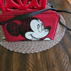 May include: Red crossbody bag featuring a black and white Mickey Mouse graphic. The bag has a black strap and zipper. The bag is rectangular and sits on a brown woven placemat.