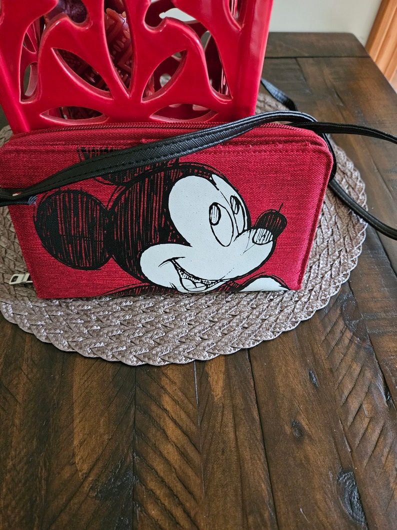 May include: Red crossbody bag featuring a black and white Mickey Mouse graphic. The bag has a black strap and zipper. The bag is rectangular and sits on a brown woven placemat.