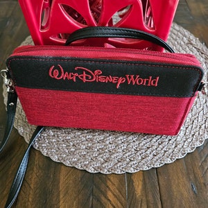 May include: Red and black wallet with the text "Walt Disney World" embroidered in red. The wallet has a black strap and zipper. The background includes a red decorative object and a brown woven placemat.