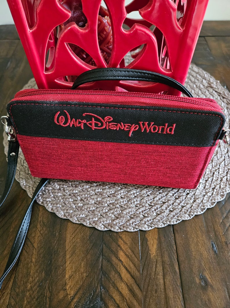 May include: Red and black wallet with the text "Walt Disney World" embroidered in red. The wallet has a black strap and zipper. The background includes a red decorative object and a brown woven placemat.