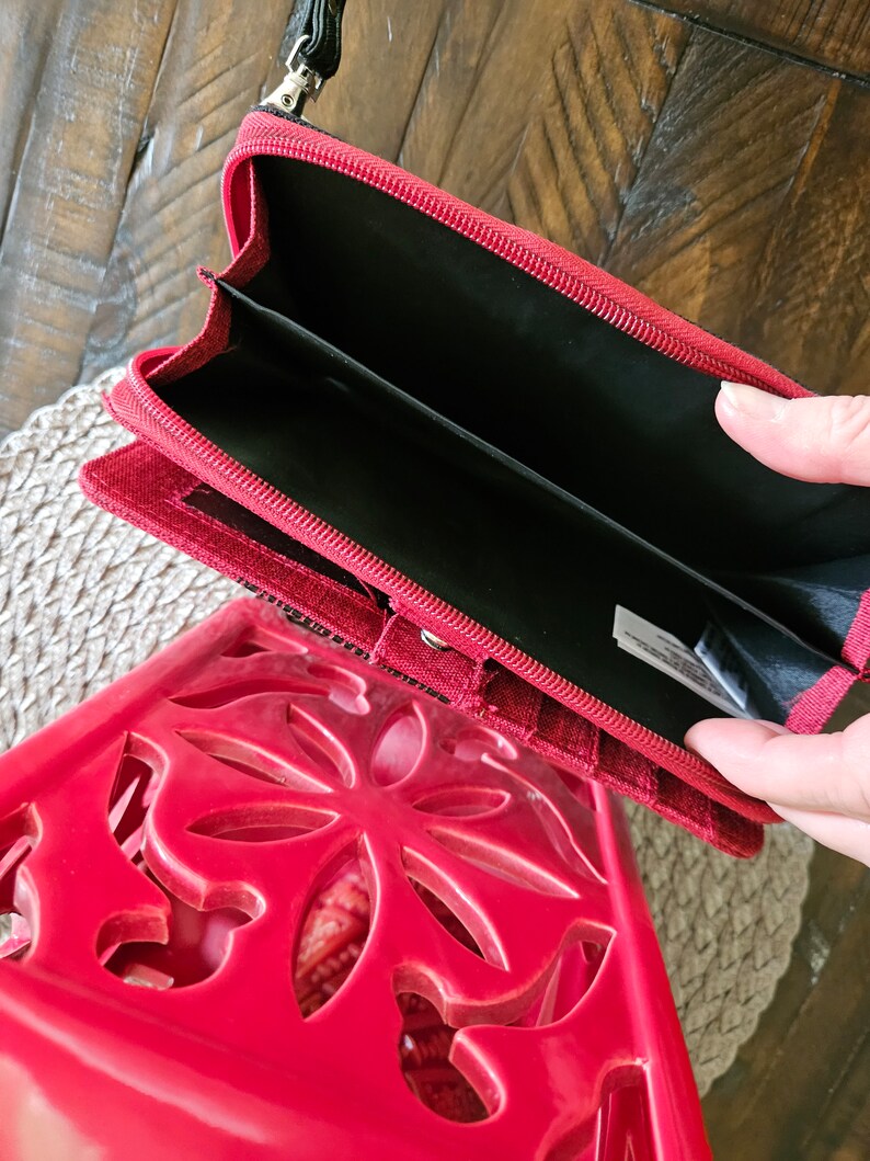 May include: Open red wallet with a black interior and a wrist strap. The wallet is held open, revealing multiple compartments. A red decorative object with a floral design is in the foreground.