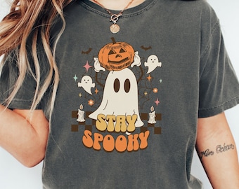 May include: A dark gray t-shirt with a white ghost wearing a jack-o-lantern on its head. The ghost is surrounded by bats, stars, and flowers. The text "Stay Spooky" is written in a retro font below the ghost.
