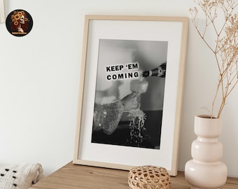 Peut inclure: Impression photographique en noir et blanc dans un cadre en bois clair. La photo montre une main versant du champagne dans un verre avec le texte "KEEP 'EM COMING" imprimé sur l'image.
