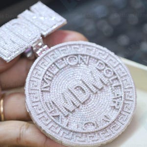 Könnte beinhalten: Ein silberner Anhänger mit einer großen, runden, diamantbesetzten Münze mit den Worten "American Dollar" und "MDLX" in der Mitte. Der Anhänger ist an einem kleineren, rechteckigen Anhänger mit dem Wort "MDLX" in Diamanten befestigt.