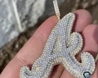 May include: A diamond-encrusted pendant in the shape of the letter "A". The pendant is primarily silver with a gold bail. The letter is covered in small, sparkling stones. The pendant is held in a hand.