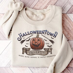 May include: A beige sweatshirt with a Halloween-themed graphic. The graphic features a pumpkin with a spooky face, bats, a broom, and the text "Halloweentown University est. 1998 Where being normal is vastly overrated".