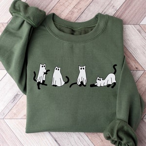 May include: A green sweatshirt featuring a white graphic of four cats dressed as ghosts. The cats are in various poses, including one cat with its arms raised in the air.