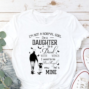 May include: White t-shirt with black text that reads "I'm not a normal girl - I'm a daughter to a Dad with wings. I used to be his angel, now he's mine."  The text is accompanied by a black silhouette of a father and daughter walking.