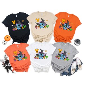 May include: Five different colored t-shirts featuring a blue Stitch character dressed as a witch with a black hat and holding a cauldron. The shirts are orange, black, white, gray, and beige.