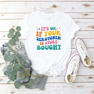Puede incluir: Camiseta blanca con un gr&aacute;fico colorido que dice "Est&aacute; bien si tu serotonina se compra en la tienda".