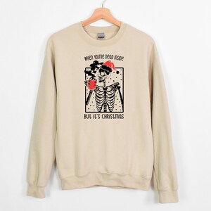 May include: A beige sweatshirt with a black and white illustration of a skeleton wearing a Santa hat and holding a mug. The text "When you're dead inside but it's Christmas" is printed on the sweatshirt.