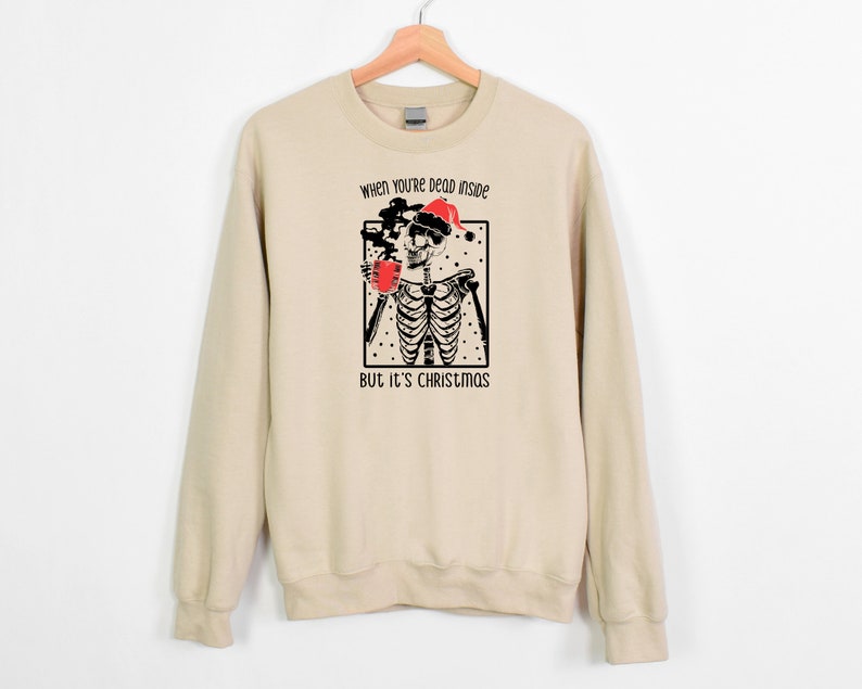 May include: A beige sweatshirt with a black and white illustration of a skeleton wearing a Santa hat and holding a mug. The text "When you're dead inside but it's Christmas" is printed on the sweatshirt.