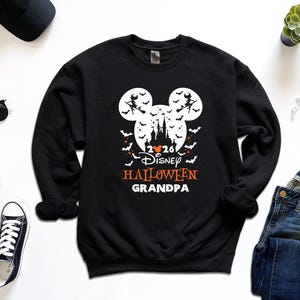 May include: A black long-sleeve sweatshirt featuring a white graphic of a castle silhouette inside a Mickey Mouse head shape, surrounded by bats and witches. The text reads 2026, Disney, HALLOWEEN, and GRANDPA in white and orange lettering.