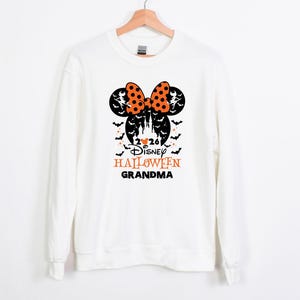 May include: A white long-sleeve sweatshirt featuring a black and orange graphic of Mickey Mouse ears with a polka dot bow, a castle silhouette, bats, and the text 2026 Disney HALLOWEEN GRANDMA printed in black and orange on the front centre.