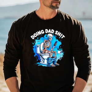May include: Black long-sleeve shirt with a humorous graphic. The graphic features a skeleton sitting on a toilet, using a phone, with the text "DOING DAD SHIT" in white. The design includes blue accents.