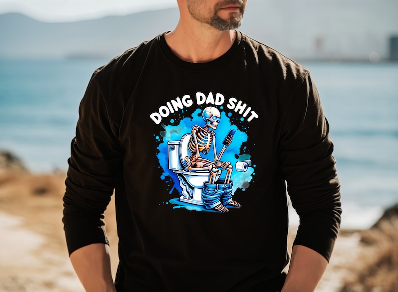 May include: Black long-sleeve shirt with a humorous graphic. The graphic features a skeleton sitting on a toilet, using a phone, with the text "DOING DAD SHIT" in white. The design includes blue accents.