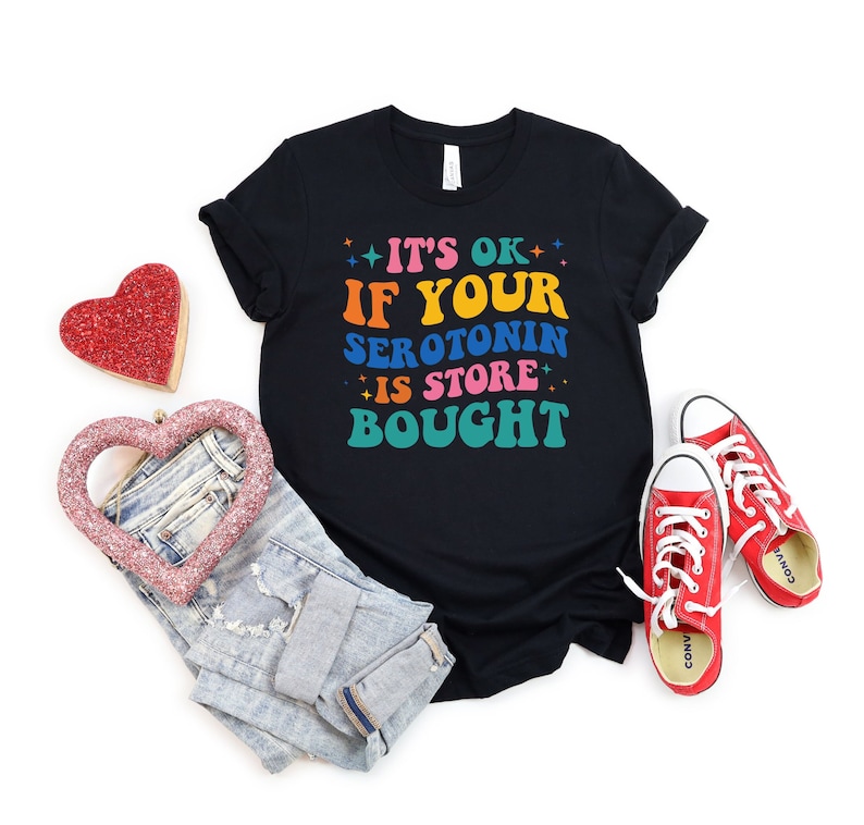 Puede incluir: Camiseta negra con el texto "It's OK if your serotonin is store bought" en colores arco&iacute;ris. La camiseta se combina con jeans azules y zapatillas rojas.