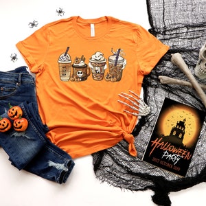 Pu&ograve; includere: Maglietta arancione con una grafica di quattro bevande al caff&egrave; a tema Halloween. La maglietta &egrave; appoggiata su una superficie bianca con un paio di jeans blu, un teschio e un invito a una festa di Halloween.