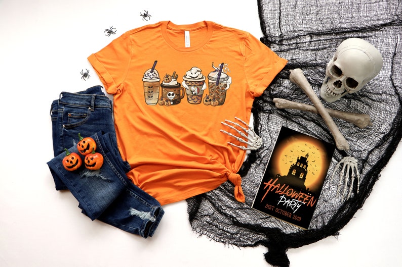 Pu&ograve; includere: Maglietta arancione con una grafica di quattro bevande al caff&egrave; a tema Halloween. La maglietta &egrave; appoggiata su una superficie bianca con un paio di jeans blu, un teschio e un invito a una festa di Halloween.