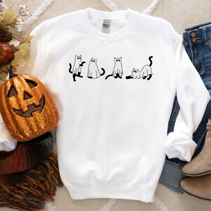 May include: White sweatshirt featuring a black and white illustration of four cats dressed as ghosts. The sweatshirt is lying on a white surface with a pumpkin, a blanket, jeans, and boots.