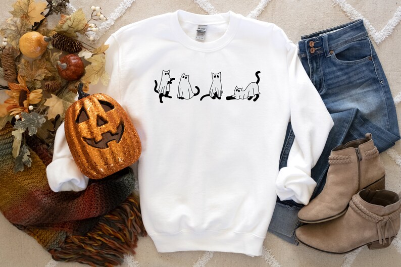 May include: White sweatshirt featuring a black and white illustration of four cats dressed as ghosts. The sweatshirt is lying on a white surface with a pumpkin, a blanket, jeans, and boots.