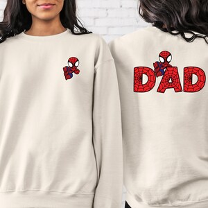 May include: Two cream-coloured sweatshirts with a cartoon Spiderman design. One sweatshirt has a small Spiderman on the front and the other has a larger Spiderman on the back with the word "DAD" spelled out in red and black.