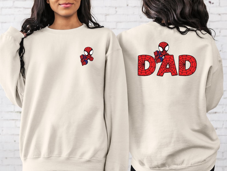May include: Two cream-coloured sweatshirts with a cartoon Spiderman design. One sweatshirt has a small Spiderman on the front and the other has a larger Spiderman on the back with the word "DAD" spelled out in red and black.