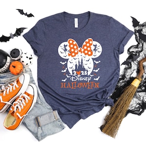 May include: A heather navy blue t-shirt featuring a white graphic of a castle silhouette inside a Mickey Mouse head shape, topped with an orange polka-dot bow. The design includes flying bats, the text 2025 DISNEY HALLOWEEN, and two witch figures.