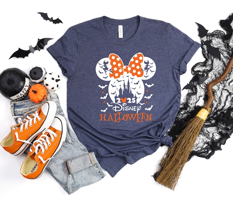 May include: A heather navy blue t-shirt featuring a white graphic of a castle silhouette inside a Mickey Mouse head shape, topped with an orange polka-dot bow. The design includes flying bats, the text 2025 DISNEY HALLOWEEN, and two witch figures.