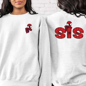 May include: Two white sweatshirts featuring a red and blue Spiderman graphic. One sweatshirt has the word "SIS" in red with a spiderweb design.