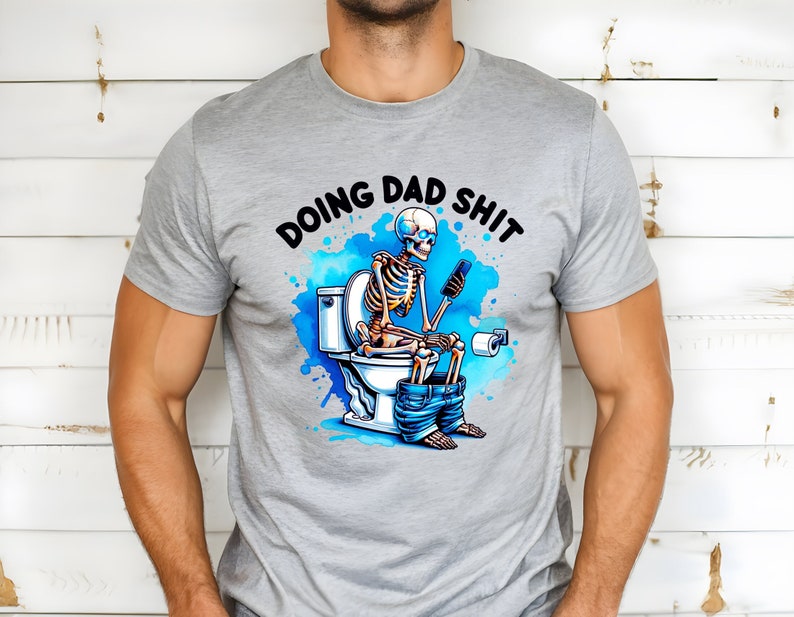 May include: A gray t-shirt with a humorous graphic. The design features a skeleton sitting on a toilet, using a phone, with the words "DOING DAD SHIT" above. The graphic includes blue watercolor-style accents.