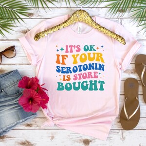 Puede incluir: Camiseta rosa con el texto "It's ok if your serotonin is store bought" en colores arco&iacute;ris.