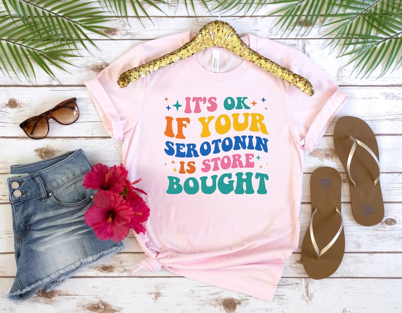 Puede incluir: Camiseta rosa con el texto "It's ok if your serotonin is store bought" en colores arco&iacute;ris.